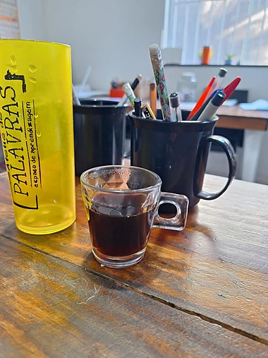 Xícara de café ao lado de um copo personalizado com a marca Ateliê de Palavras, sobre a mesa de trabalho