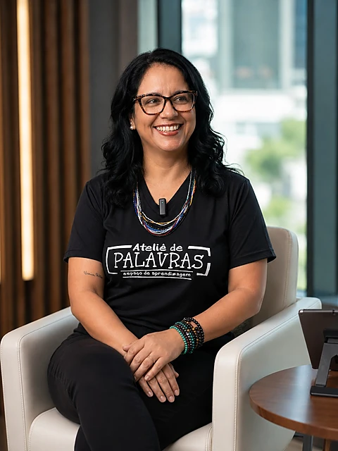 Professora Bruna Valério sorrindo, sentada em poltrona, vestindo camiseta preta com a estampa Ateliê de Palavras — espaço de aprendizagem